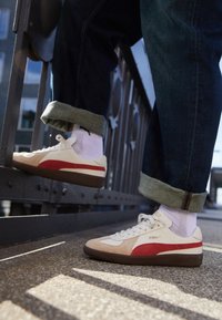 Sneakers Puma s bílým koženým svrškem, červeným pruhem, akcenty z hnědého semiše, bílými šňůrkami a hnědými gumovými podrážkami, nošeny s tmavě modrými džíny.