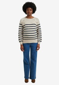 Cremfarbener und marineblauer gestreifter Pullover mit Rundhalsausschnitt, drei Knöpfen an einer Schulter, getragen zu blauen weit geschnittenen Jeans und braunen Absatzschuhen.