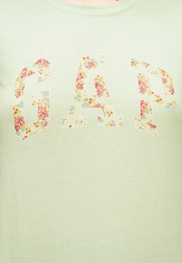 Λογότυπο GAP με μεγάλα γράμματα γεμάτα πολύχρωμο λουλουδάτο σχέδιο σε ανοιχτό πράσινο ύφασμα.