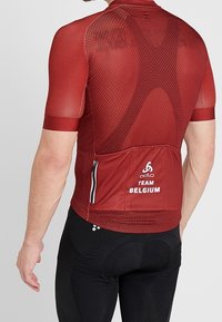 Rød cykeltrøje med meshtekstur, korte ærmer og en lynlåslomme på ryggen. Har 'Team Belgium' logo og kontrasterende syninger.