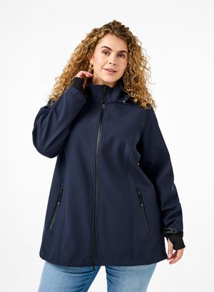 Frau mit lockigem Haar, die eine marineblaue Jacke mit Reißverschluss, Reißverschlusstaschen und Daumenlöchern an den Bündchen trägt, posiert vor einem weißen Hintergrund.