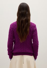 Person mit langen, welligen braunen Haaren in einem lila, kabelgestrickten Pullover und einem beigen Rock, die von hinten vor einem schlichten, hellen Hintergrund steht.