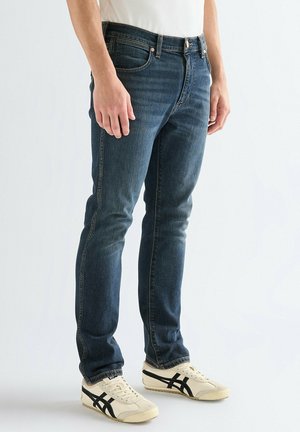 Persona con jeans ajustados azul oscuro y zapatillas blancas con rayas negras, de pie contra un fondo claro y liso.