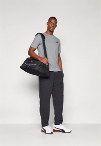 Mladenič v sivi majici, črnih hlačah, belih športnih copatih, nosi črno športno torbo prek rame in stoji pred enobarvno ozadjem.