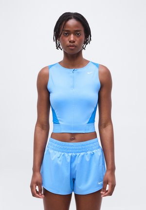 Jonge vrouw staand, gekleed in een lichtblauw mouwloos sportcrop top en bijpassende korte broek tegen een effen witte achtergrond.