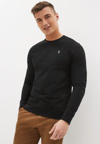 Svart bomullströja med långa ärmar, rund halsringning, och en liten broderad logga på bröstet. Har ribbad nederkant och muddar.
