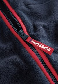 Tamsiai mėlynos spalvos fliso audinys su raudona užtrauktuku, turintis prekės ženklo raudoną traukimo kilpą. Tekstūra yra minkšta, o užtrauktukas apjuostas kontrastine apdaila.