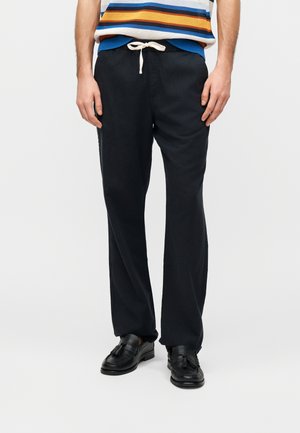 Férfi fekete zsinóros nadrágban, fekete bojtos mokaszinban és kék, narancs, fehér csíkos ujjatlan pulóverben.