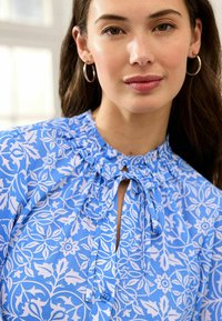 Blusa floreale blu con scollatura arricciata. Presenta motivi bianchi intricati, un'apertura a chiave e bottoni frontali. Tessuto morbido al tatto.