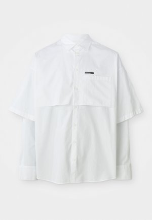 Camisa branca de botão de manga curta com bolso peitoral e rótulo da marca. Feita de um material leve de algodão com uma textura suave.