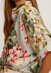 Blouse à motif floral en satin gris clair avec des fleurs rose vif et jaune, des feuilles vertes et des manches retroussées. Col boutonné.
