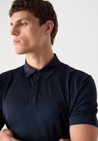 Camisa polo azul marinho com acabamento brilhante, colarinho proeminente e fecho de botão. Apresenta um logótipo bordado discreto no peito.