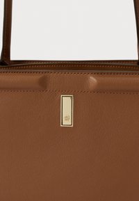 Bolsa de mão de couro castanho com chapa retangular dourada e alças estruturadas visíveis na parte superior.