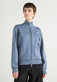 Blue zip-up sweatshirt made of soft fabric, featuring a high collar, two side pockets, and a subtle logo on the chest. Smooth texture.