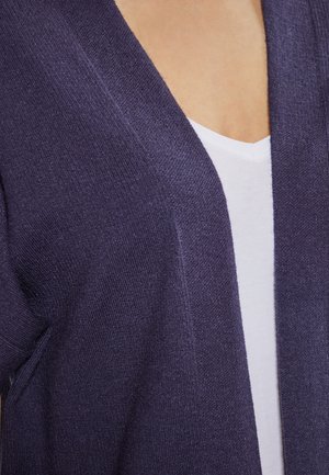 Cardigan tricotat violet cu decolteu adânc în V, având o textură moale și un design cu față deschisă, purtat peste un tricou alb cu decolteu în V.