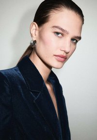 Joven mujer con el cabello peinado hacia atrás que lleva pendientes de aro de plata y un blazer de terciopelo azul oscuro, mirando ligeramente de lado.