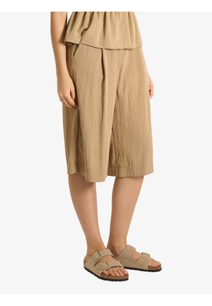 Donna che indossa pantaloni beige larghi fino al ginocchio e sandali beige con doppia fibbia, in piedi su sfondo bianco.