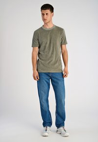 Uomo in piedi che indossa una camicia a maniche corte verde oliva, jeans blu e sneakers bianche su uno sfondo chiaro e semplice.
