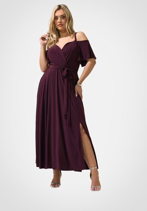 Kobieta ubrana w długość do ziemi, bordową suknię z odkrytymi ramionami, wiązaną w talii i z rozcięciem z boku, w połączeniu ze srebrnymi sandałami na wysokim obcasie i bransoletką.