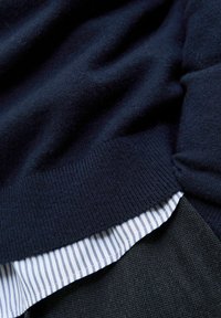 Suéter de lana navy con puños acanalados, superpuesto a una camisa a rayas blanca y navy, combinado con una tela texturizada gris por debajo.