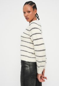 Pull rayé blanc avec des lignes grises, texture tricotée, col rond, manches longues, porté avec un pantalon en cuir noir. Fond uni.