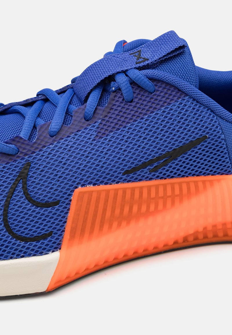 Chaussure de sport bleue avec une tige en maille, présentant un logo noir et une semelle texturée orange. Le design comprend des lacets plats et un col rembourré.