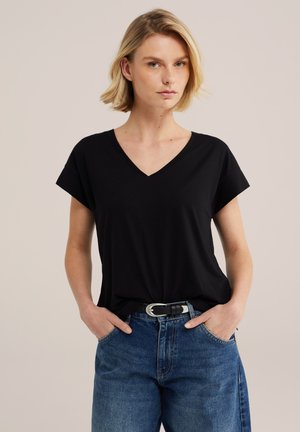 Femme aux cheveux blonds courts portant un t-shirt noir à col en V rentré dans un jean bleu avec une ceinture noire, debout les mains dans les poches.