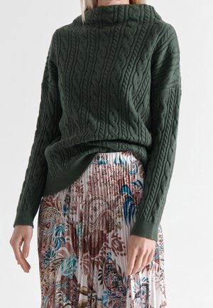 Woman wearing dark green cable-knit sweater and pleated skirt with brown, blue, and beige floral paisley patterns, shown from neck to mid-thigh.