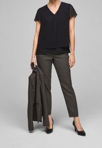 Blusa nera a strati con maniche corte, scollo a V; pantaloni con motivo, in mano una giacca abbinata. Un paio di scarpe nere con tacco alto.