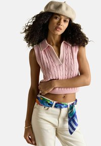 Rózsaszín kötött ujjatlan felső bordázott szegéllyel, V-nyakkal és fehér szegéllyel; fehér farmerrel és több színű mintás sállal párosítva.