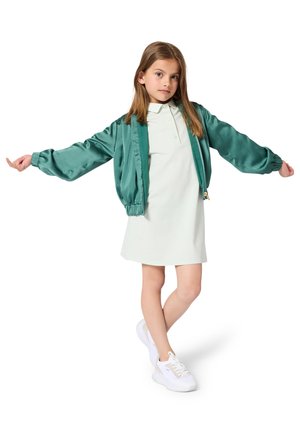 Jeune fille portant une robe légère, une veste en satin teal avec les bras tendus, et des baskets blanches, se tenant devant un fond blanc.