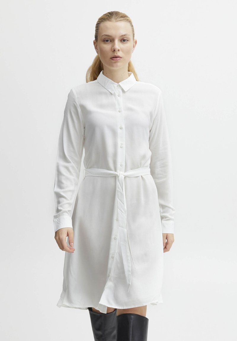 Robe blanche à manches longues et boutons, en tissu lisse, avec un col, une ceinture à la taille et un ourlet au genou. Design simple et épuré.