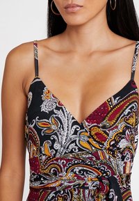 Robe noire avec un motif cachemire en rouge, jaune et blanc. Présente des bretelles fines, un décolleté en V profond et un détail de taille nouée.