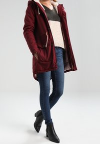 Parka burdeos con capucha forrada de piel sintética y dos bolsillos delanteros, combinada con un suéter de rayas grises y cremas, junto con vaqueros ajustados y botines negros.