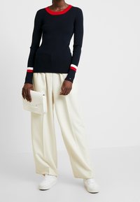 Osoba v černém svetru s dlouhým rukávem a červenobílými manžetami, širokých krémových kalhotách, bílých teniskách a držící bílou kabelku.