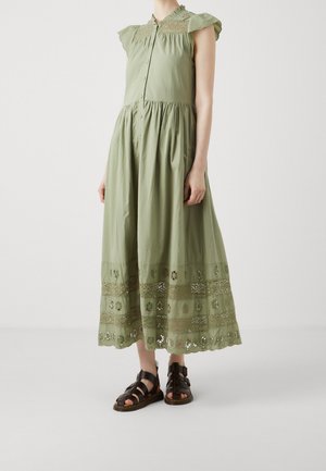 Woman wearing a sleeveless olive green midi dress with lace details and button-up front, paired with black strapped sandals.