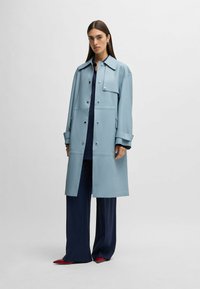 Light blue, long, waterproof coat with a classic collar, snap buttons, and side pockets; worn over a navy outfit and red shoes.
