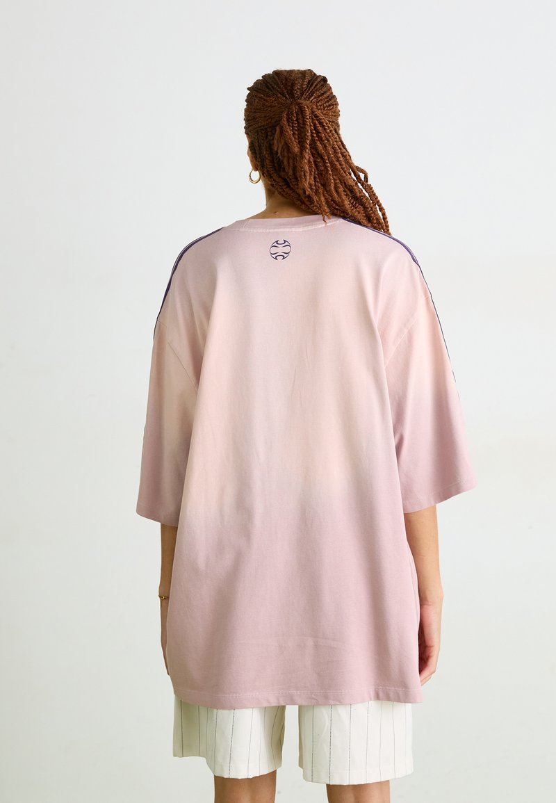 Przytulny, oversize różowy t-shirt z krótkim rękawem, z okrągłym logo na plecach i kontrastowymi akcentami w kolorze granatowym na ramionach.