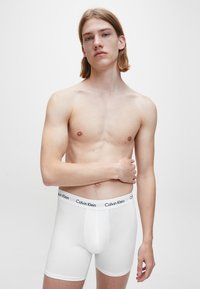 Mladic s dugom kosom stoji naslonjen na jednostavnu sivu pozadinu, odjeven u bijele Calvin Klein bokserice, s jednom rukom prekriženom preko tijela.