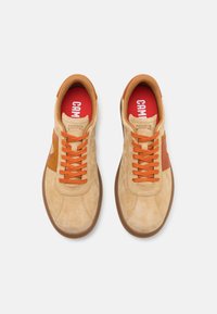 Sneaker in suede beige con suola in gomma marrone, con lacci arancioni e un accento marrone. Soletta rossa con il marchio "CAMPER" in bianco.