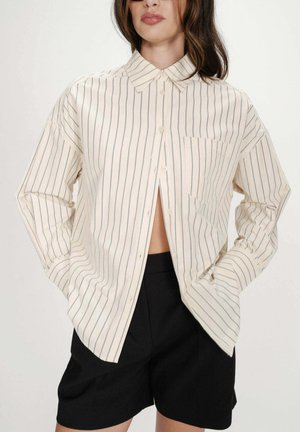 Femme portant une chemise crème surdimensionnée à fines rayures verticales noires, déboutonnée en bas, associée à un short noir, les mains dans les poches.