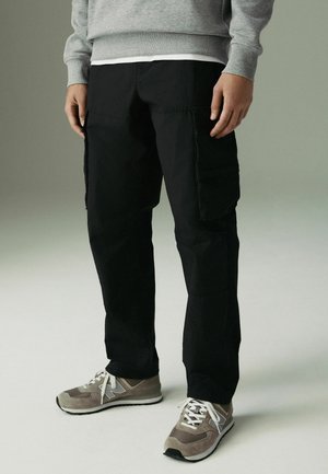 Black cargo pants with multiple side pockets, made from durable fabric. Paired with gray athletic shoes featuring white laces and logo accents.