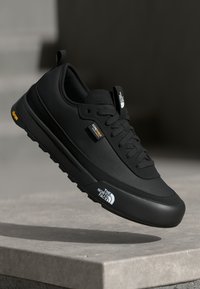 Black sneaker with a mesh upper and rubber sole. Features include a cushioned tongue, prominent branding, and a textured surface.