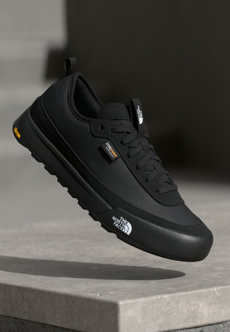 Black sneaker with a mesh upper and rubber sole. Features include a cushioned tongue, prominent branding, and a textured surface.