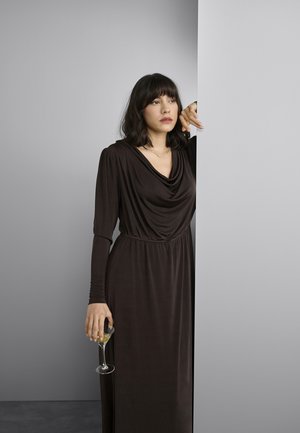 Rochie maro, cu mâneci lungi, cu un decolteu drapat și talie strânsă, realizată dintr-un material neted. Femeie ținând un pahar de cocktail, stând în picioare lângă un perete.