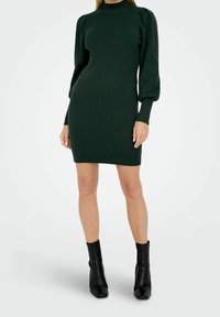 Femme portant une robe en maille côtelée vert foncé avec des manches longues ballon et des bottines noires, debout devant un fond blanc.