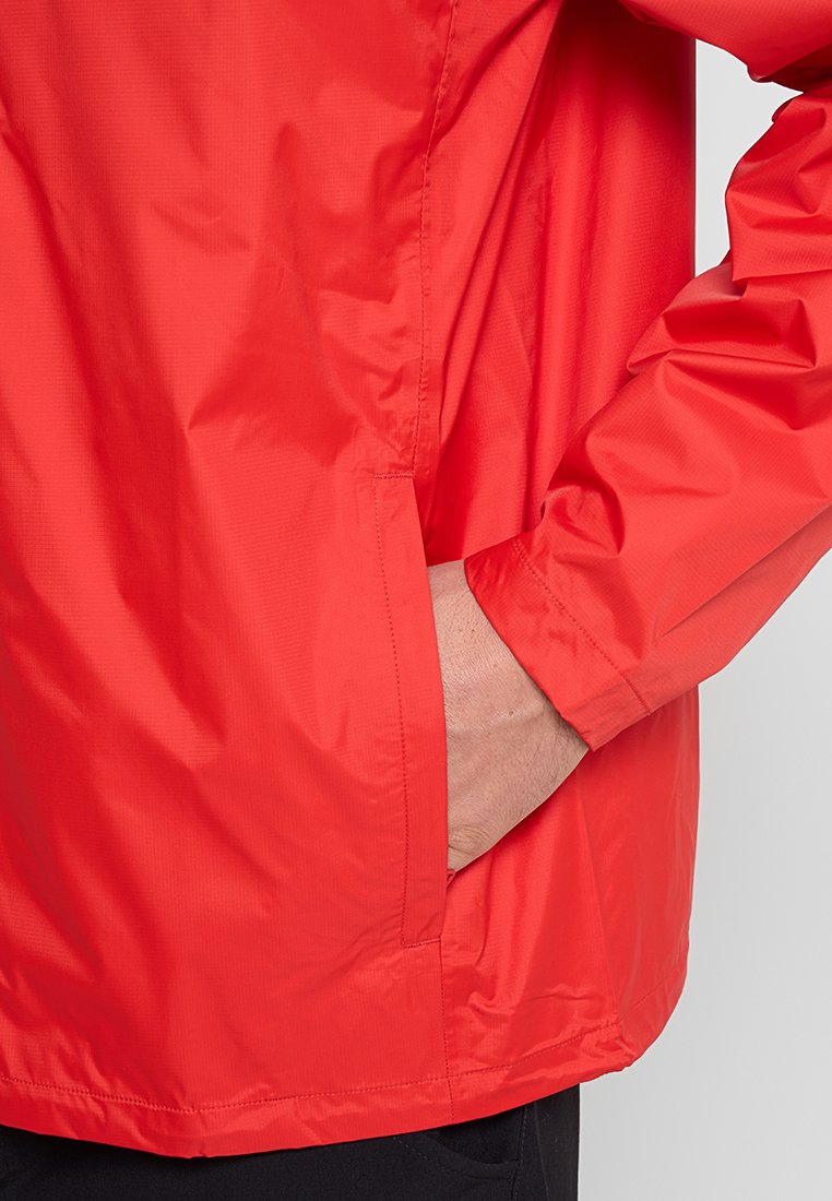 Mano in tasca di un vivace giubbotto impermeabile a maniche lunghe di colore rosso brillante, indossato sopra pantaloni neri, su uno sfondo chiaro.