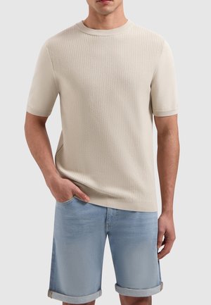 Beige gebreide trui met korte mouwen en een gestructureerd ontwerp, ronde halslijn en een iets getailleerde vorm, gecombineerd met lichtblauwe broekjes met omslag.