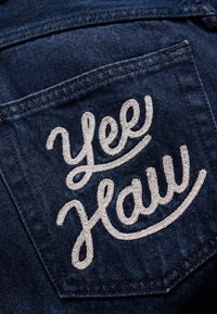 Tummansininen farkkutakin tasku, jossa on valkoisella brodeerattu teksti "Yee Haw" käärmefontilla. Kangas on klassista laatua.