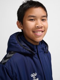 Jonge jongen lacht, draagt een marineblauw capuchonjack met een sportlogo op de borst en patroon schouderstrepen.